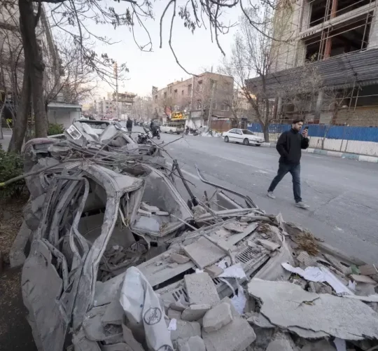іранець йде по дорозі біля зруйнованих будівель та автівок