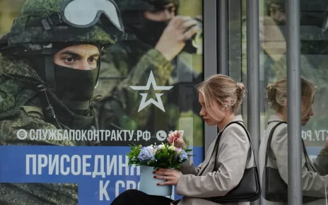 росіянка сидить на зупинці з плакатом на якому заклик до приєднання до військової служби