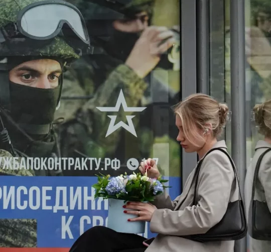 росіянка сидить на зупинці з плакатом на якому заклик до приєднання до військової служби