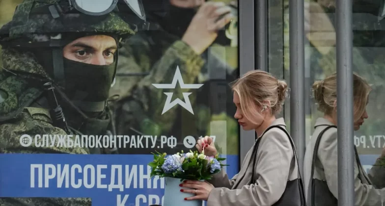 росіянка сидить на зупинці з плакатом на якому заклик до приєднання до військової служби