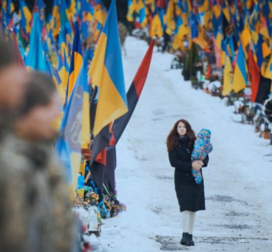 молода жінка йде з малюком на руках повз могили загиблих українських воїнів
