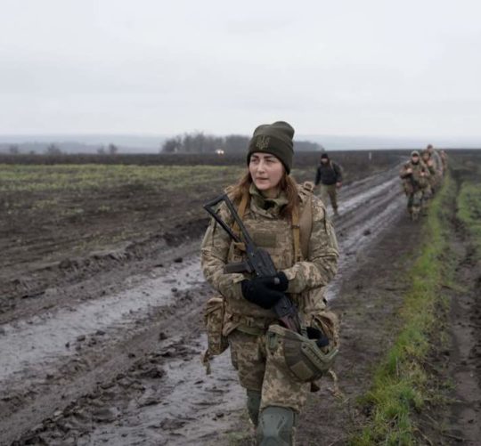 українська жінка військова йде по переду з автоматом по українському полі