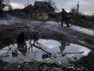 Чоловік веде велосипед по грунтовій морозній дорозі: на передньому плані — велика калюжа, а на задньому — зруйнований будинок