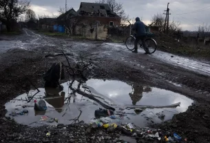 Чоловік веде велосипед по грунтовій морозній дорозі: на передньому плані — велика калюжа, а на задньому — зруйнований будинок