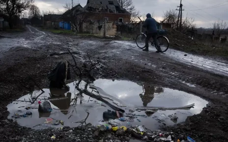 Чоловік веде велосипед по грунтовій морозній дорозі: на передньому плані — велика калюжа, а на задньому — зруйнований будинок