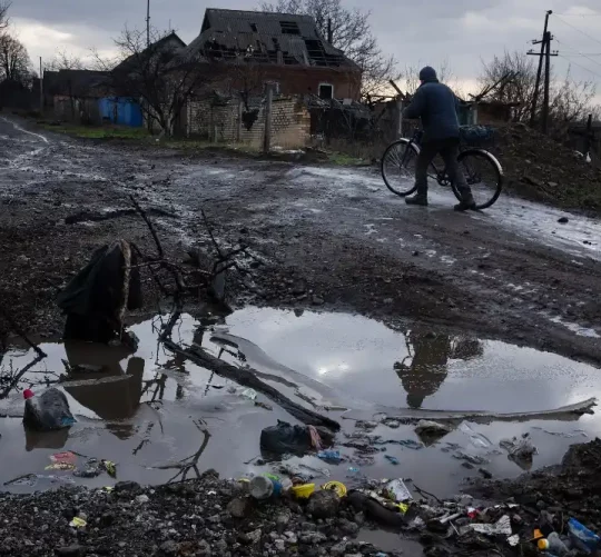 Чоловік веде велосипед по грунтовій морозній дорозі: на передньому плані — велика калюжа, а на задньому — зруйнований будинок