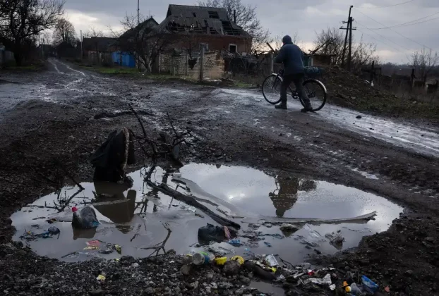 Чоловік веде велосипед по грунтовій морозній дорозі: на передньому плані — велика калюжа, а на задньому — зруйнований будинок