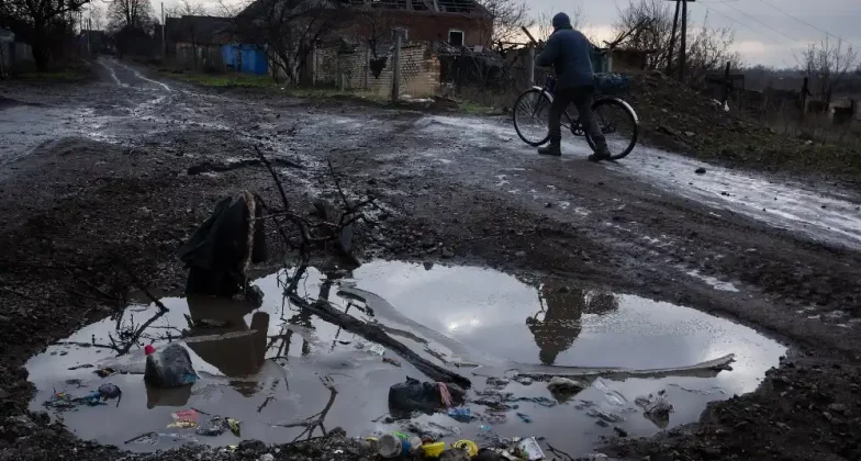 Чоловік веде велосипед по грунтовій морозній дорозі: на передньому плані — велика калюжа, а на задньому — зруйнований будинок