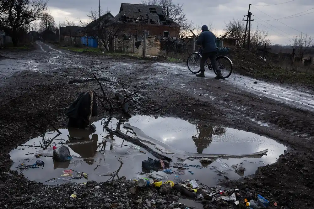 Чоловік веде велосипед по грунтовій морозній дорозі: на передньому плані — велика калюжа, а на задньому — зруйнований будинок