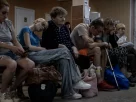 На лавці сидить кілька людей, деякі з них схилили голови або опустили їх. Одна людина тримає тростину, а перед нею на землі лежать сумки