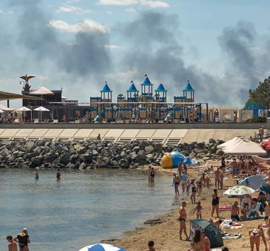Місцеві жителі та гості міста Одеса відпочивають на пляжі, коли піднімається дим від пожежі, що спалахнула внаслідок російського ракетного удару 29 липня