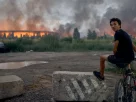 Підліток сидить на велосипеді й спостерігає, як полум’я здіймається з палаючого логістичного складу після удару російської балістичної ракети по Києву