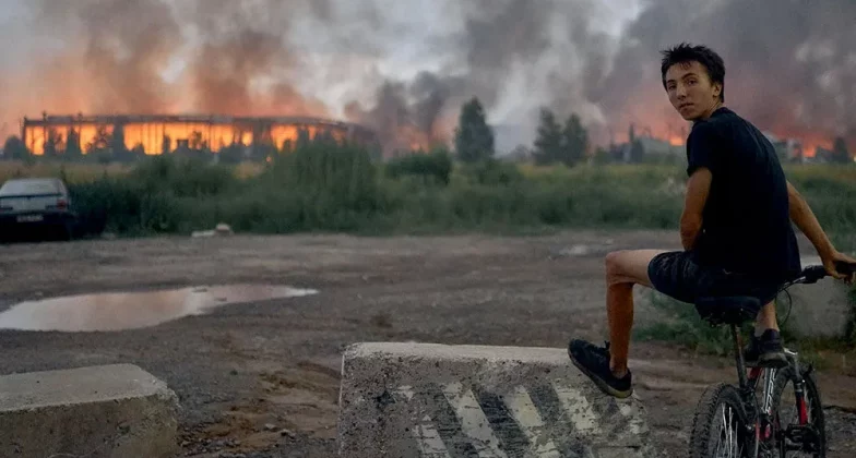 Підліток сидить на велосипеді й спостерігає, як полум’я здіймається з палаючого логістичного складу після удару російської балістичної ракети по Києву